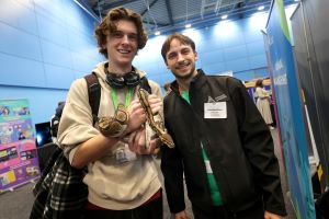 Student in a brown hoodie holds a snake while smiling at the camera. He is next to another man in a black jacket.