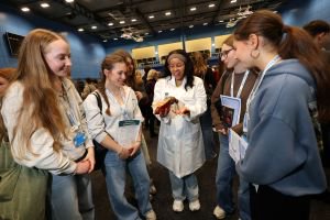 A grioup of young people stand around a woman in a white lab coat holding an animal. They look engaged and smiling.